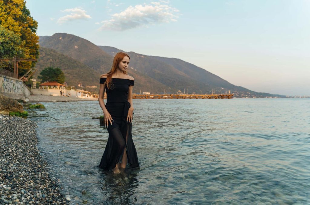 Artystyczna sesja kobieca w plenerze – ruda kobieta w długiej czarnej sukni stojąca w wodzie na kamienistej plaży, w tle wysokie góry i jezioro, fotografia artystyczna Śląsk.
