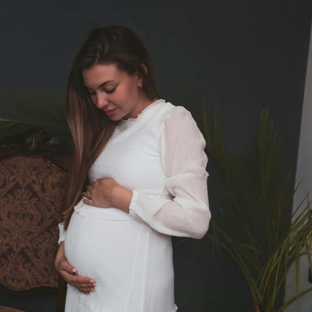 Elegancka sesja ciążowa w studio Katowice – przyszła mama w białej zwiewnej sukni trzymająca się za brzuszek, ciemne tło i rośliny doniczkowe, fotografia brzuszkowa Śląsk.