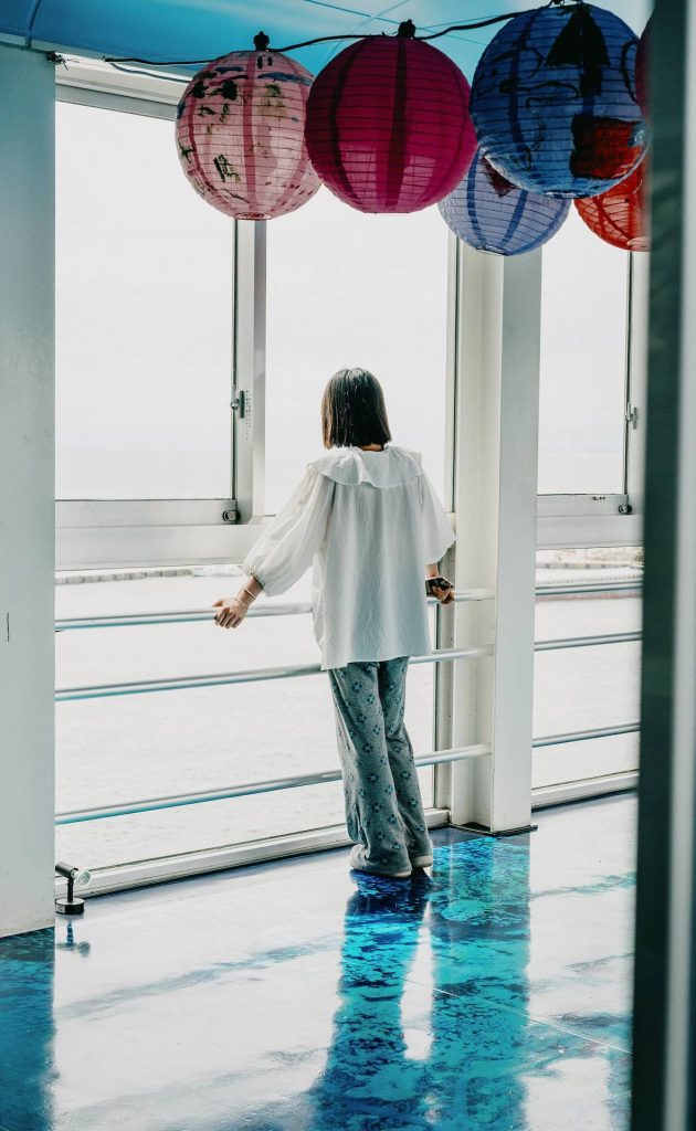 Nostalgiczna sesja kobieca w plenerze – dziewczyna w białej bluzce patrząca przez okno na wodę, kolorowe lampiony pod sufitem, fotografia lifestyle Katowice.