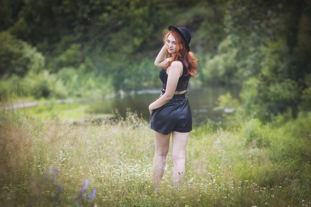 Naturalna sesja kobieca w plenerze – ruda modelka w czarnym kapeluszu i letniej stylizacji na ukwieconej łące, w tle las i rzeka, fotografia portretowa Katowice.