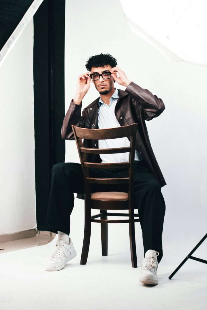 Męska sesja portretowa w studio w Katowicach – model w brązowej skórzanej kurtce i okularach siedzący tyłem na krześle, minimalistyczne tło, fotografia wizerunkowa Śląsk.