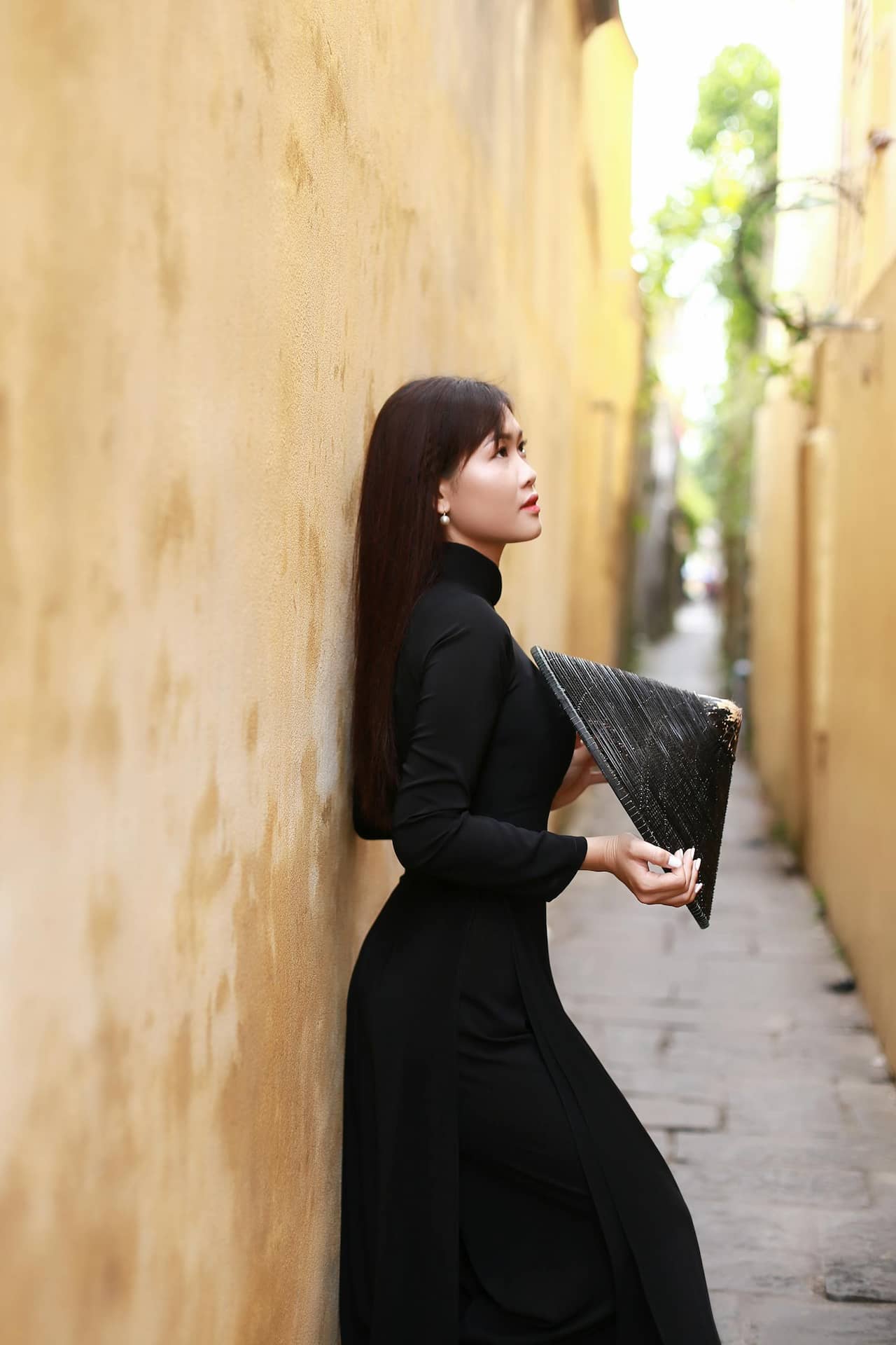 Elegancka sesja kobieca w Katowicach – młoda kobieta w długiej czarnej sukni z tradycyjnym kapeluszem, pozująca w wąskiej, żółtej uliczce, fotografia lifestyle Śląsk.