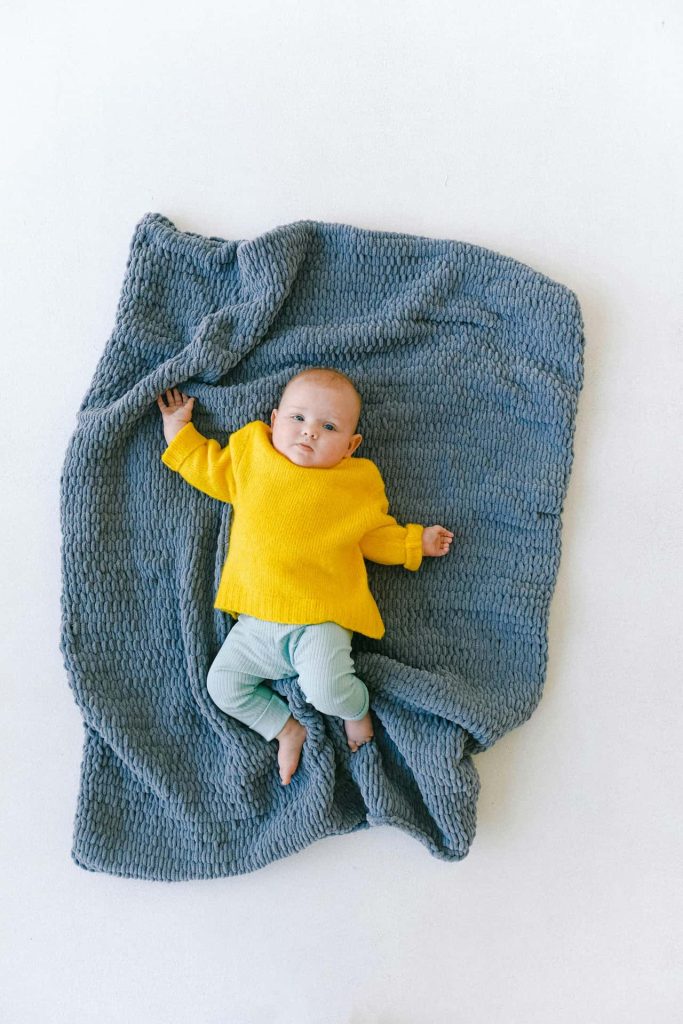 Minimalistyczna sesja noworodkowa w Katowicach – niemowlę w jaskrawym żółtym sweterku leżące na grubym, niebieskim plecionym kocu, fotografia dziecięca studio Śląsk.