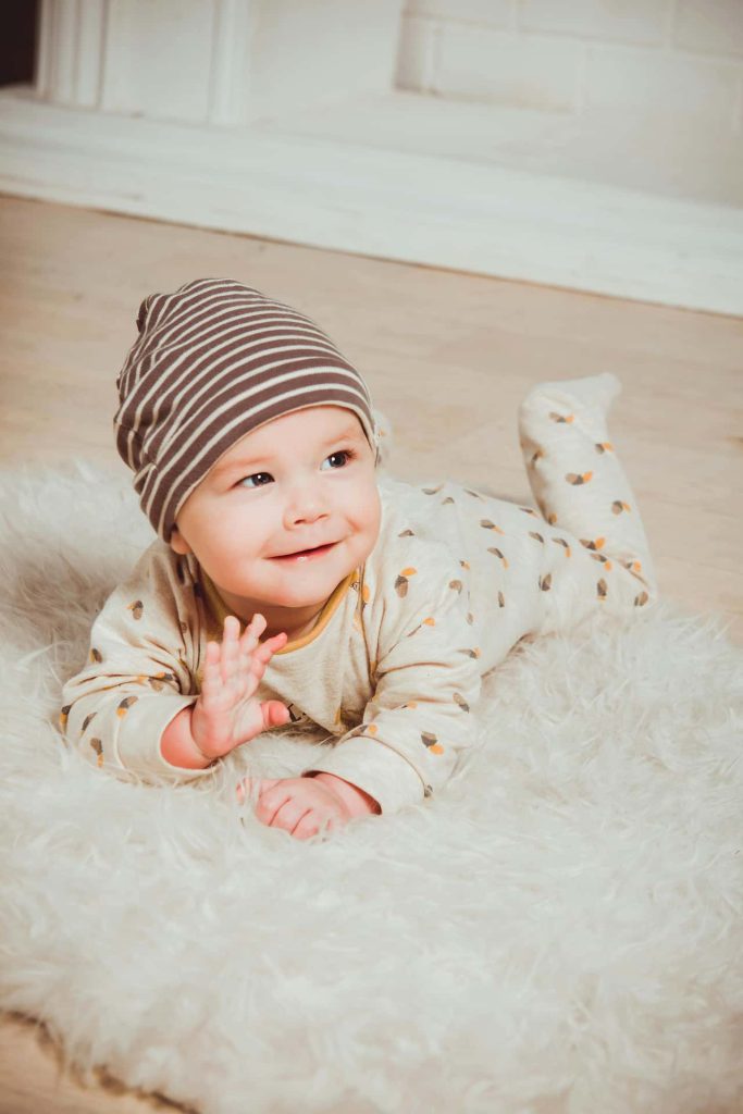 Radosna sesja niemowlęca w Katowicach – uśmiechnięte niemowlę w pasiastej brązowej czapce leżące na białym puszystym dywanie, naturalna fotografia dziecięca studio Śląsk.