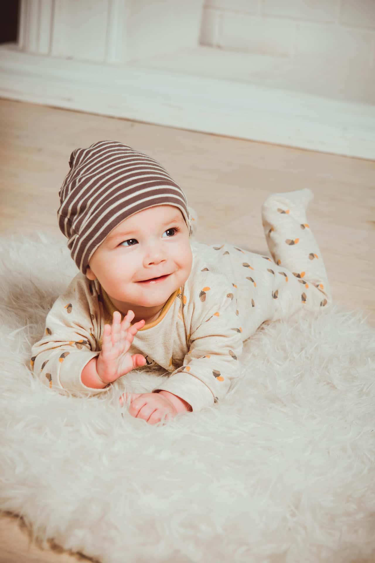Radosna sesja niemowlęca w Katowicach – uśmiechnięte niemowlę w pasiastej brązowej czapce leżące na białym puszystym dywanie, naturalna fotografia dziecięca studio Śląsk.