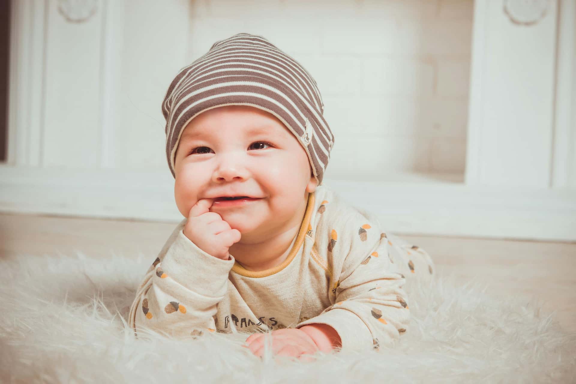 Wesołe sesje noworodkowe Katowice – uśmiechnięte niemowlę w prążkowanej czapce leżące na białym puszystym dywanie, jasne studio fotograficzne Śląsk.