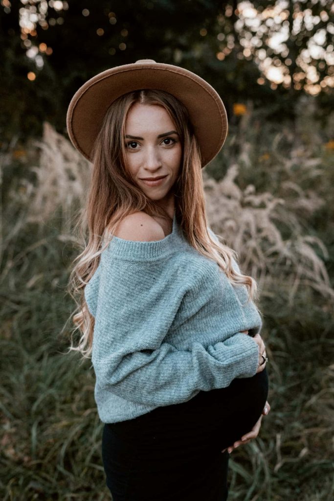 Sesja ciążowa w stylu boho Katowice – przyszła mama w brązowym kapeluszu i szarym swetrze trzymająca się za brzuszek na tle wysokich traw, jesienna fotografia brzuszkowa.