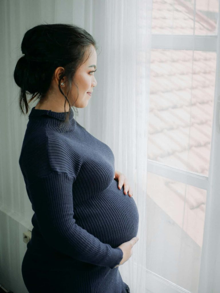 Naturalna sesja ciążowa w domu Katowice – kobieta w ciąży w granatowym golfie patrząca przez okno, miękkie światło dzienne, fotografia brzuszkowa lifestyle Śląsk.