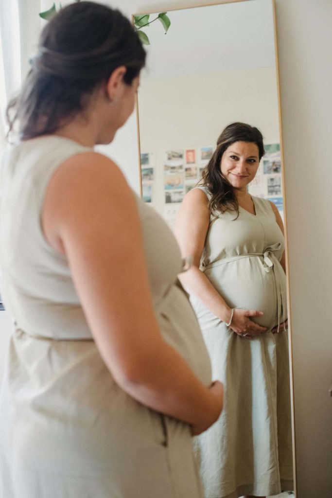 Naturalna sesja ciążowa Katowice – przyszła mama w beżowej sukience uśmiechająca się do swojego odbicia w lustrze, domowa fotografia brzuszkowa Śląsk.