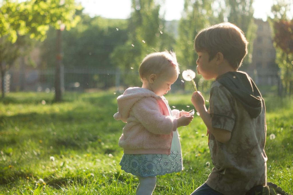 Naturalna sesja dziecięca w plenerze Katowice – mała dziewczynka w różowej bluzie i chłopiec dmuchający latawce z mlecza, rodzeństwo bawiące się w ogrodzie o zachodzie słońca, fotografia dziecięca Śląsk.