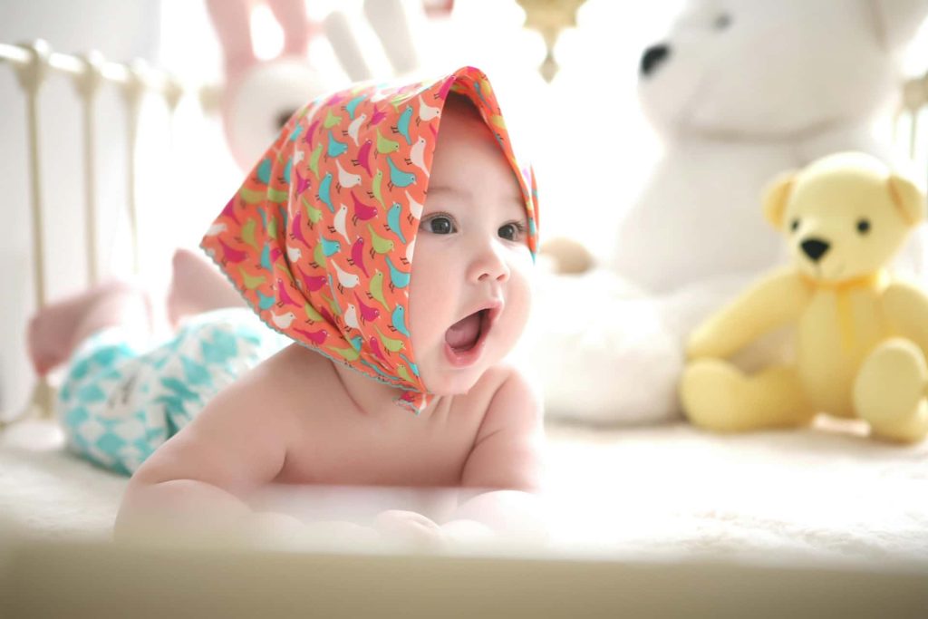 Kolorowa sesja dziecięca w Katowicach – niemowlę w pomarańczowej chustce w ptaszki leżące na brzuszku, w tle biały i żółty miś, jasna fotografia lifestyle Śląsk.