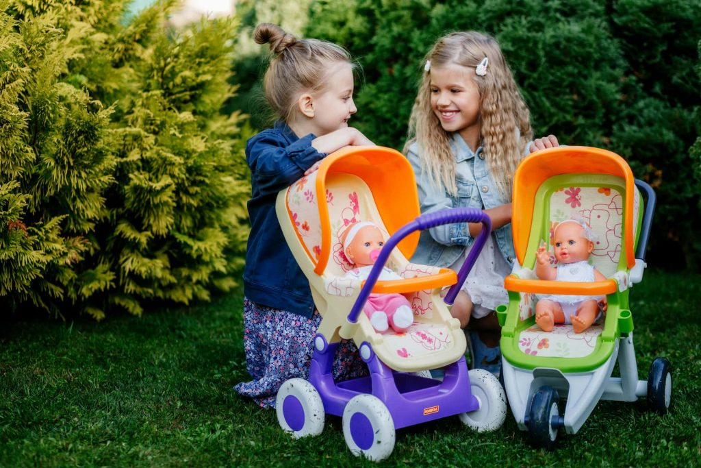 Naturalna sesja dziecięca Katowice – dwie uśmiechnięte dziewczynki bawiące się wózkami z lalkami na trawie, fotografia lifestyle rodzeństwa w ogrodzie, Śląsk.